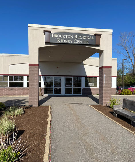 Entrance of Brockton Regional Kidney Center on a clear day, showing a modern medical facility with glass doors, clean landscaping, and clear signage