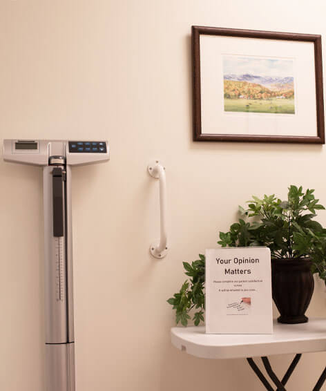 Medical clinic interior with a digital physician beam scale, wall-mounted grab bar, framed landscape painting, potted green plant on a small white table, and a sign that reads 'Your Opinion Matters' for patient feedback collection